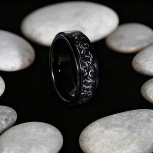 May include: A black ring with a detailed pattern around the band. The ring is set against a black background and surrounded by white stones. The ring's design is intricate and appears to be etched or engraved.