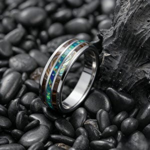 May include: A silver ring with a band featuring a central inlay of iridescent blue and green abalone shell, flanked by bands of dark wood and lighter-colored material. The ring is set against a background of dark gray stones.