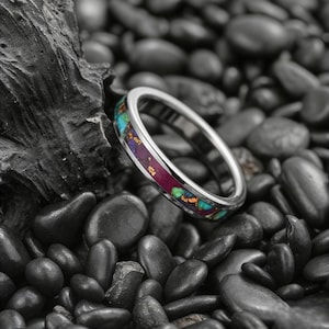 Könnte beinhalten: Ein silberner Ring mit einem farbenfrohen Inlay aus türkisfarbenen, lila und goldfarbenen Steinen. Der Ring liegt vor einem Hintergrund aus schwarzen Kieselsteinen und einem dunklen, strukturierten Felsen.