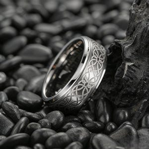 May include: A silver-colored ring with an intricate Celtic knot design engraved around the band. The ring is set against a backdrop of dark, smooth stones, highlighting its polished surface and detailed pattern. The ring's design is symmetrical and detailed.