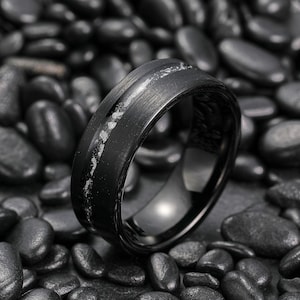 May include: A black ring with a brushed metal finish and a central inlay of white speckled material. The ring is set against a backdrop of dark, smooth stones, creating a contrast in textures and tones. The ring's design is modern and sleek.