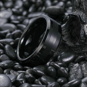 以下が含まれることがあります： 黒色のリング。つや消し加工された金属仕上げと研磨された内側が特徴です。リングは、暗く滑らかな石の背景に配置されています。リングのデザインは、テクスチャパターンの中央バンドとシルバーカラーのエッジを備えています。