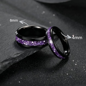 May include: Two black rings with purple gemstone accents. One ring is 8mm wide, the other is 4mm wide. The gemstones are set in a band around the rings. The rings are displayed on a dark, textured surface.