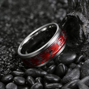 May include: A silver-colored ring with a red band featuring a repeating spider design. The ring is set against a dark, textured background of small, black stones and a larger rock formation.