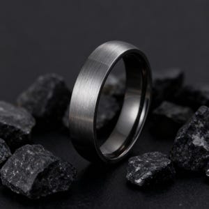 May include: A dark gray ring with a brushed metal finish. The ring is set against a backdrop of dark gray rocks, creating a contrast that highlights the ring's texture and design. The ring's band has a smooth, polished interior.