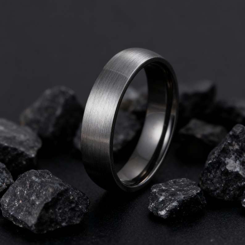 May include: A dark gray ring with a brushed metal finish. The ring is set against a backdrop of dark gray rocks, creating a contrast that highlights the ring's texture and design. The ring's band has a smooth, polished interior.