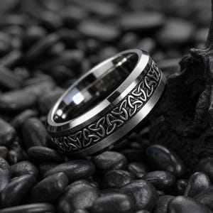 May include: A silver-toned ring with a Celtic knot design. The ring has a brushed metal finish on the band and a polished interior. The Celtic knot pattern is etched around the ring's exterior. The ring is set against a background of dark, smooth stones.