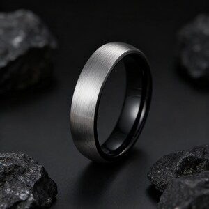 May include: A close-up of a silver-colored ring with a brushed metal finish. The ring has a black interior and is set against a dark background with dark rocks.
