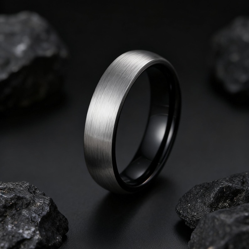 May include: A close-up of a silver-colored ring with a brushed metal finish. The ring has a black interior and is set against a dark background with dark rocks.