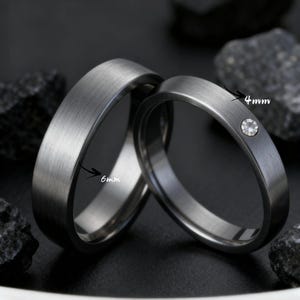 Op de afbeelding: Twee donkergrijze trouwringen. De ene band is 6 mm breed, de andere 4 mm breed en voorzien van een kleine, heldere edelsteen. Beide ringen hebben een geborstelde metalen afwerking en staan tegen een donkere achtergrond.