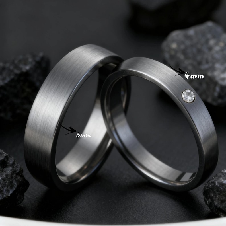May include: Two dark gray wedding bands. One band is 6mm wide, the other is 4mm wide and features a small, clear gemstone. Both rings have a brushed metal finish and are set against a dark background.
