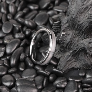 Puede incluir: Un anillo plateado con una incrustación blanca. El anillo se encuentra sobre un fondo de piedras negras lisas y una formación rocosa oscura y texturizada. La superficie pulida del anillo refleja la luz.