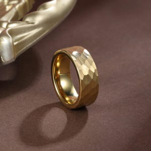 May include: A gold-colored ring with a textured, hammered surface. The ring has a wide band and a polished interior. The ring is displayed on a brown surface, with a gold-colored decorative element in the background.