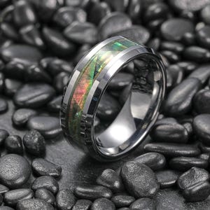 Puede incluir: Un anillo plateado con una incrustación iridiscente en tonos verdes, rosas y naranjas. El anillo destaca sobre un fondo de piedras grises oscuras. Una joya llamativa con un diseño colorido.