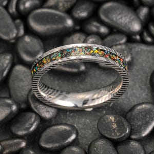 May include: A silver ring with a colorful inlay of various gemstones. The ring has a textured, patterned band and is set against a backdrop of black stones. The gemstones display a vibrant array of colors, including red, yellow, and green.