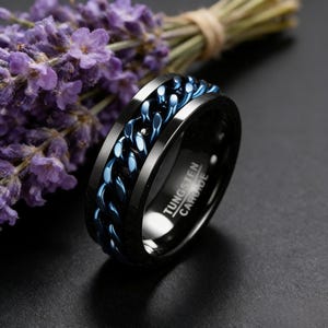 May include: Black tungsten carbide ring featuring a blue chain design. The ring is inscribed with "Tungsten Carbide". The image includes a bunch of purple flowers. The ring is a stylish accessory.