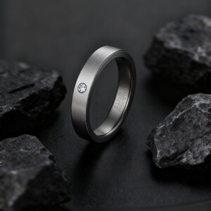 May include: A brushed silver ring with a single, small, clear gemstone. The ring has a flat band and a minimalist design. The ring is set against a dark background with dark gray rocks.