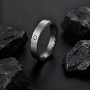 May include: A brushed silver ring with a single, small, clear gemstone. The ring has a flat band and a minimalist design. The ring is set against a dark background with dark gray rocks.
