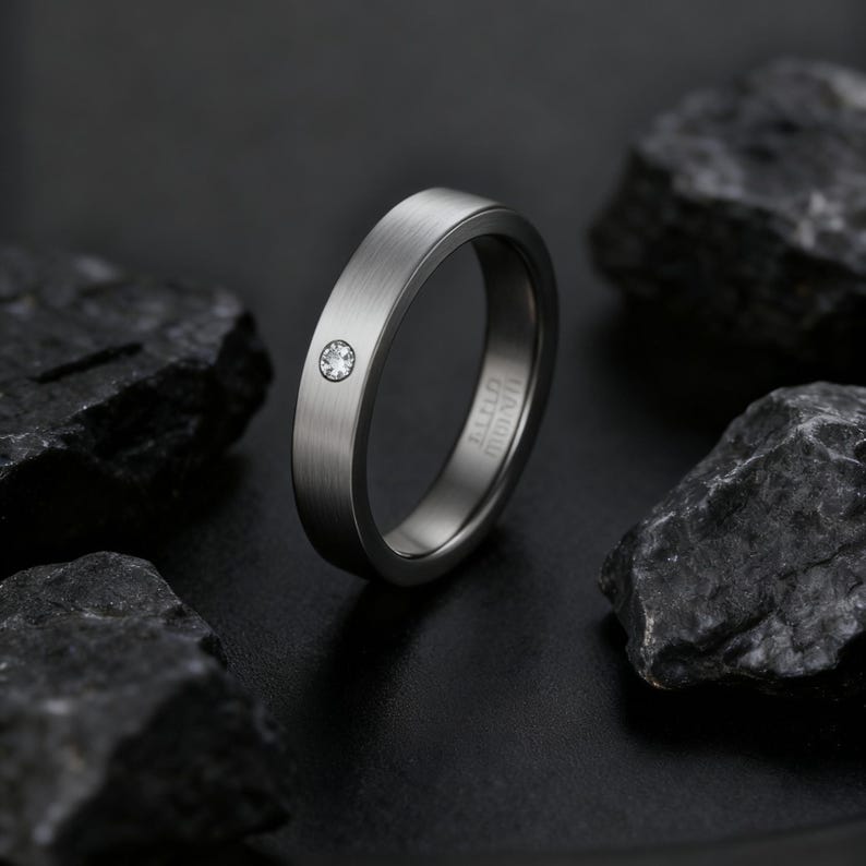 May include: A brushed silver ring with a single, small, clear gemstone. The ring has a flat band and a minimalist design. The ring is set against a dark background with dark gray rocks.