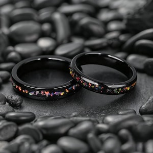 Può includere: Due anelli neri con un design di pietre preziose colorate intarsiate. Le pietre preziose presentano un mix di tonalità rosse, arancioni, gialle e viola. Gli anelli sono posizionati su uno sfondo di pietre scure e lisce.