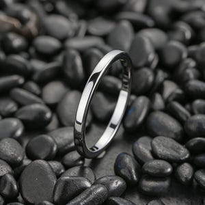 Può includere: Un anello d'argento lucido con una fascia liscia e arrotondata. L'anello è posto su uno sfondo di pietre scure, lisce e arrotondate. La superficie dell'anello riflette la luce, evidenziando la sua lucentezza metallica.