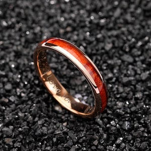 May include: Rose gold ring with a polished, reddish-brown wood inlay. The ring has a smooth, rounded profile and the words "Love You" are engraved on the inside. The ring is set against a dark, textured background.