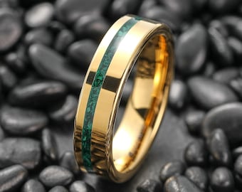 May include: A gold-colored ring with a central inlay of green stone. The ring has a polished, reflective surface, and the green inlay provides a striking contrast to the gold. The ring is set against a background of dark, smooth stones.
