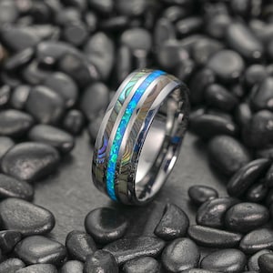 Puede incluir: Un anillo plateado con una banda central de incrustaciones de concha de abulón azul vibrante e iridiscente. El diseño del anillo presenta un acabado metálico pulido y se coloca sobre un fondo de piedras oscuras y lisas. Una joya llamativa.