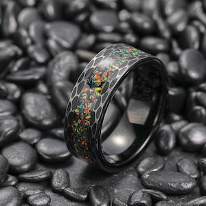Può includere: Un anello nero con un motivo a nido d'ape e un'incastonatura di opale colorata. L'opale presenta una miscela di tonalità rosse, arancioni, gialle, verdi e blu. L'anello è posto su uno sfondo di pietre scure.