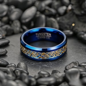 May include: A blue tungsten carbide ring with a gold Celtic knot design. The ring has the text "Tungsten Carbide" engraved on the inside. The ring is displayed on a dark surface with black stones.