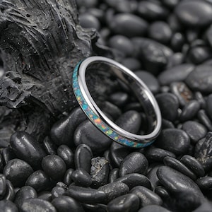 May include: A silver ring featuring a band of iridescent blue and orange opal. The ring is set against a backdrop of black stones and a dark, textured surface.