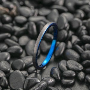 May include: A slender ring featuring a brushed grey exterior and a bright blue inner band. The ring is displayed against a background of smooth, dark grey stones. The design is minimalist and elegant, with a modern aesthetic.