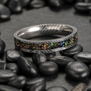 May include: A silver ring with a textured band and a colorful inlay of iridescent stones. The ring is set against a dark background with black stones.
