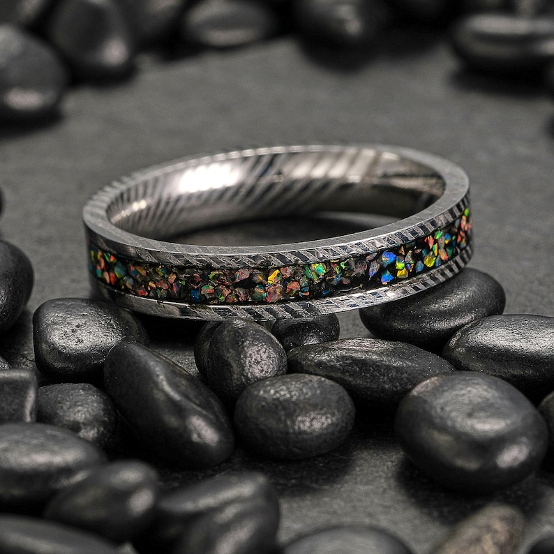 May include: A silver ring with a textured band and a colorful inlay of iridescent stones. The ring is set against a dark background with black stones.