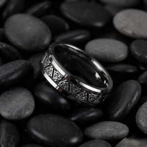 May include: A silver-colored ring with a geometric pattern around the band. The ring is set against a background of dark gray stones, creating a contrast that highlights the ring's design. The ring's surface reflects light, adding to its visual appeal.