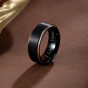 Puede incluir: Un anillo negro con un acabado de metal cepillado y una banda interior dorada. El anillo tiene el texto grabado "I Love You" en el interior. El anillo está sobre una superficie marrón.