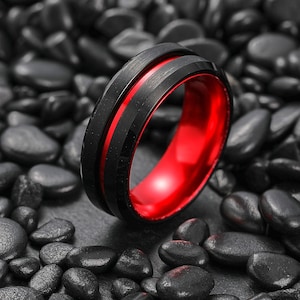 Puede incluir: Un anillo negro con interior rojo. El anillo tiene un diseño acanalado en la banda exterior. El anillo está sobre un fondo de piedras negras.