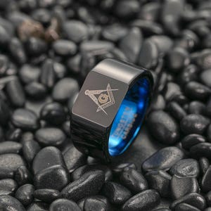 以下が含まれることがあります： 青い内側の黒いタングステンカーバイドリング。リングの前面にはフリーメーソンのシンボルが刻まれています。リングは黒い石の背景に配置されています。