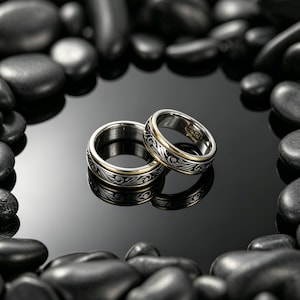 May include: Two silver rings with gold detailing and ornate black floral designs. The rings are displayed on a reflective black surface, framed by smooth, dark grey stones. The rings are likely for a couple.