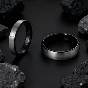 May include: Two tungsten carbide rings. One ring has a brushed silver exterior and a black interior, with a small, clear gemstone. The other ring has a brushed silver exterior and a black interior. Both rings are set against a black background with dark rocks.