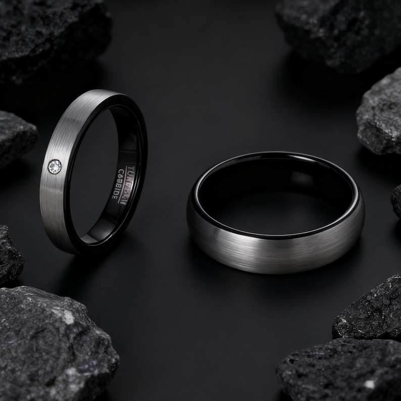 May include: Two tungsten carbide rings. One ring has a brushed silver exterior and a black interior, with a small, clear gemstone. The other ring has a brushed silver exterior and a black interior. Both rings are set against a black background with dark rocks.