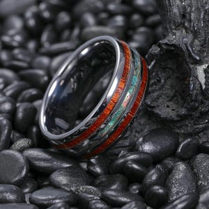Pode incluir: Um anel prateado com acabamento polido. O anel apresenta uma faixa de madeira castanho-avermelhada e material verde e azul iridescente. O anel está sobre um fundo de pedras pretas.