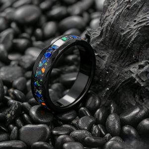 May include: A black ring with a faceted band and a colorful inlay of blue, green, and orange stones. The ring is set against a backdrop of black stones and a dark, textured surface.