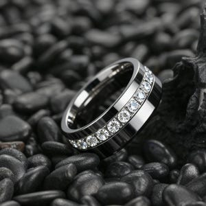 Puede incluir: Un anillo plateado con dos filas de piedras claras y brillantes. El anillo se muestra sobre un fondo de guijarros oscuros y lisos, creando un contraste llamativo.