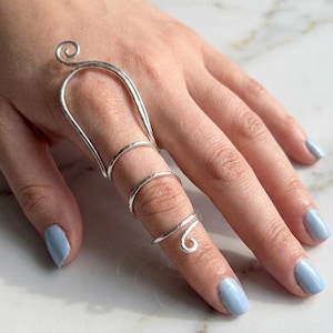 May include: A silver-colored finger ring with a unique design. The ring features a curved bar that extends over the top of the finger, connected to three bands that wrap around the finger. The ring has a spiral detail at the tip.