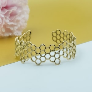 May include: A gold-colored cuff bracelet with a honeycomb design. The openwork bracelet is made of interconnected hexagonal shapes. The background is a gradient of yellow and white, with a pink flower in the upper left corner.