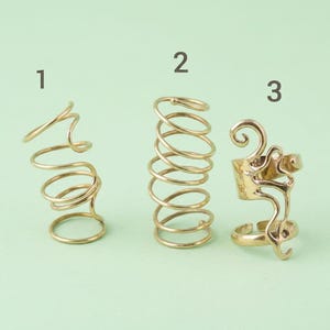 May include: Three gold-colored rings are displayed against a light green background. Ring 1 is a spiral design, ring 2 is a coil, and ring 3 has an ornate, abstract design. The rings are made of a shiny metal.
