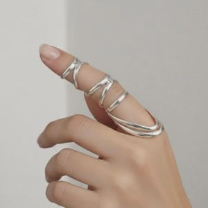 May include: A silver-colored ring wraps around the finger with multiple bands and a unique design. The ring has a modern, minimalist style, and is shown on a hand against a neutral background. The ring's design is eye-catching and elegant.