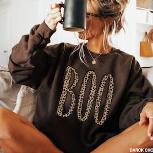 May include: A dark chocolate brown crewneck sweatshirt featuring the word BOO in large, leopard print applique letters across the chest. The garment has long sleeves and a relaxed fit, with the text DARCK CHOCOLATE printed in the bottom corner.