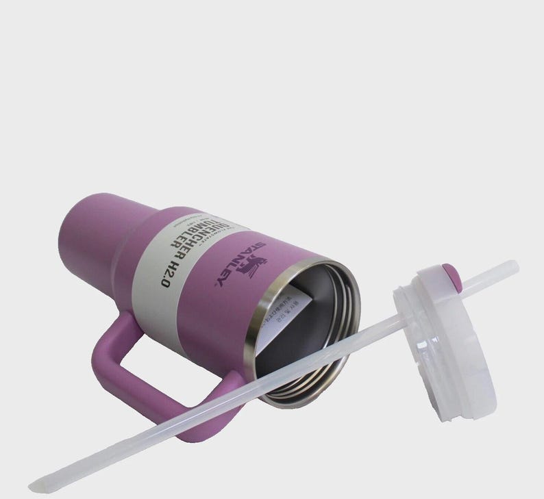 May include: A purple Stanley Quencher H2.0 tumbler with a handle, a white band, and a clear straw. The lid is white with a purple button. The Stanley logo is visible on the side of the tumbler.